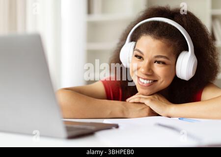 Lächelnde Schwarze Frau, Die Kabellose Kopfhörer Mit Laptop Trägt Und Sich Am Schreibtisch Beugt Stockfoto