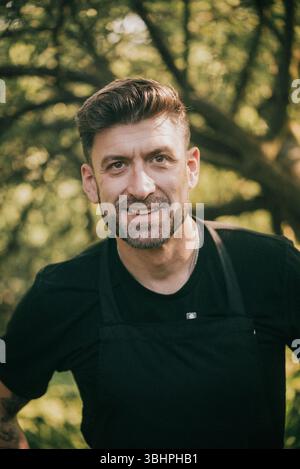 Ein Mann mit Bart und schwarzem Hemd lächelt. Er trägt eine Schürze. Der Mann steht in einem Wald Stockfoto
