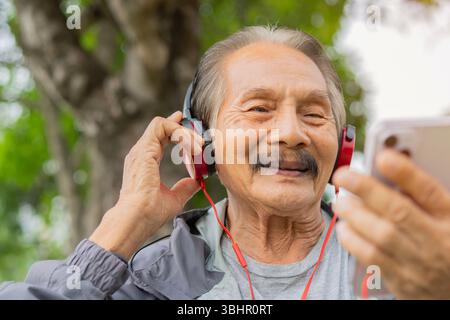 Glücklicher älterer asiatischer Mann im grünen Park, der Kopfhörer trägt und Musik hört. Ein lustiges Konzept von einem jungen Mann. Stockfoto