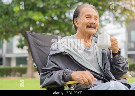 Glücklicher lächelnder älterer asiatischer Mann, der sich im Sommer morgens im Freien entspannt und eine Tasse Kaffee hält. Lebensstil-Konzept im Ruhestand. Stockfoto