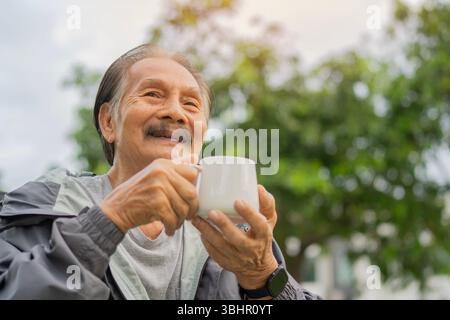 Porträt eines älteren asiatischen Mannes, der im Herbst draußen sitzt und eine Tasse Kaffee oder Tee mit Kopierraum hält. Lebensstil-Konzept im Ruhestand. Stockfoto