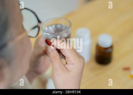 Eine ältere Frau sitzt an einem Tisch mit einem Glas Wasser und einer Flasche Pillen und nimmt sie. Rückansicht. Senioren-Gesundes-Konzept. Stockfoto