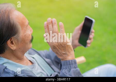 Fröhlicher älterer Mann, der im Urlaub in der Natur auf einem Stuhl sitzt und mit dem Smartphone Videoanrufe mit der Familie anstellt. Rückansicht und selektiver Fokus. Stockfoto