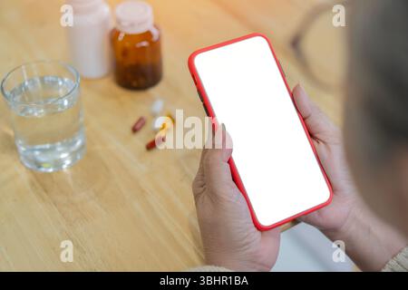 Frau mittleren Alters mit grauem Haar, die ein Handy mit leerem Bildschirm hält, in der Nähe eine Flasche mit Medikamenten auf dem Tisch. Stockfoto