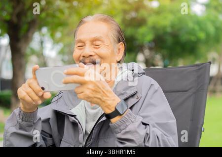 Lächelnder, aktiver Mann, der auf dem Campingstuhl sitzt und auf das Smartphone über dem Hintergrund der Natur blickt. Lebensstil-Konzept im Ruhestand. Stockfoto