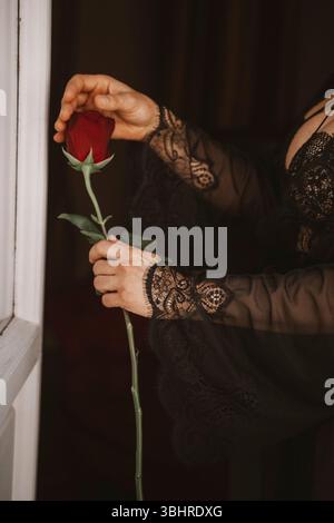 Die Frauenhand im Spitzenärmel hält sanft eine einzelne rote Rose an einem offenen Fenster. Die zarte Spitze steht im Kontrast zur leuchtenden Rose, die auf Themen von hindeutet Stockfoto