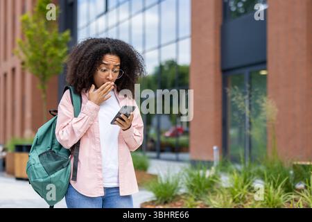 Eine junge Frau mit Brille reagiert schockiert, während sie draußen in der Nähe eines Gebäudes auf ihr Telefon blickt. Stockfoto