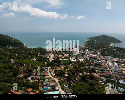 Grün bewaldete Gegend mit einem kleinen Dorf umgeben von Hügeln und einer hellen Küste in der Ferne. Ko Pha Ngan, Thailand. Stockfoto