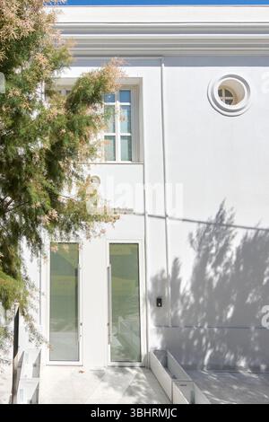 Die unberührte weiße Fassade eines modernen Gebäudes auf Rhodos mit großen Glastüren und einem eleganten runden Fenster, teilweise von einem lebhaften Fenster verdeckt Stockfoto