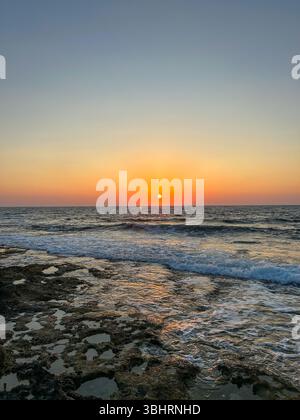 Goldener Sonnenuntergang über dem Mittelmeer mit Wellen, die sanft auf ein felsiges Ufer in Zypern krachen und eine ruhige und lebendige Szene schaffen. Stockfoto