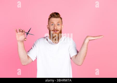 Fröhlicher bärtiger Mann in lässigem weißem T-Shirt mit Schere, Gesten auf rosa Hintergrund, verspielter Ausdruck Stockfoto