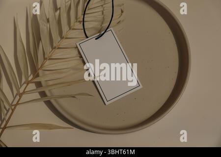 Minimalistischer flacher Lay. Leeres Geschenkanhänger, Aufdruck auf beigefarbener Keramikplatte, verziert mit getrockneten Palmblättern. Neutrale Töne, weiche Schatten. Natürlicher Sommer Stockfoto