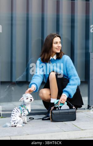 Glückliche junge Frau, die ihren Hund streichelt und hockt, vor einem modernen Glasgebäude Stockfoto