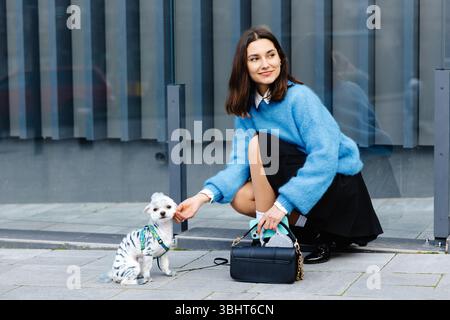 Glückliche junge Frau, die ihren Hund streichelt und hockt, vor einem modernen Glasgebäude Stockfoto