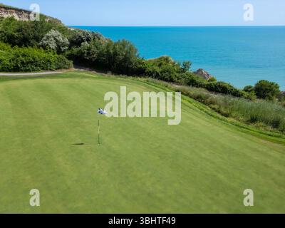 Aus nächster Nähe auf ein Golfgrün mit einer karierten Flagge in der Nähe einer malerischen Küstenklippe, umgeben von üppigem Grün und mit Blick auf ruhiges Türkis Stockfoto