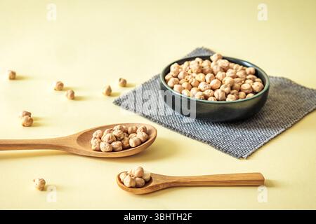 Kichererbsen in Holzlöffeln und Keramikschale auf weichgelbem Hintergrund. Gesunde Ernährung und vegetarisches Kochkonzept Stockfoto