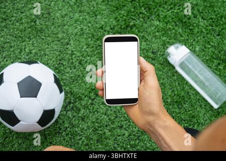 Die Hand des Fußballfans hält das Telefon mit weißem, leerem Bildschirm neben dem Fußball auf grünem Gras. Online. Moderne Technologie mit traditionellem Sport. Platz für Stockfoto