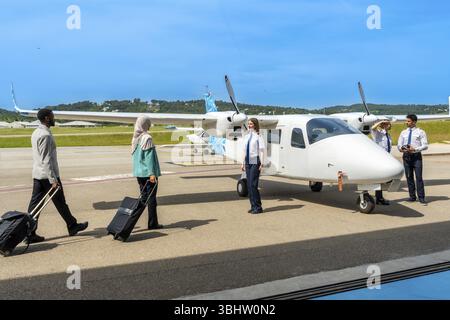 Passagiere mit Trolley-Taschen gehen in Richtung eines kleinen Flugzeugs und einer Flugbesatzung, bereit für das Einsteigen an einem sonnigen Tag am Flughafen Stockfoto