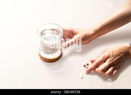 Hand hält ein Glas Wasser neben mehreren Pillen auf heller Oberfläche. Gesundheits- und Wellness-Konzepte. Stockfoto