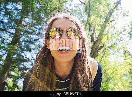 Lächelnde Frau mit langen Dreadlocks und Sonnenbrille, genießen Sie einen sonnigen Tag in einem grünen Park umgeben von Bäumen. Stockfoto