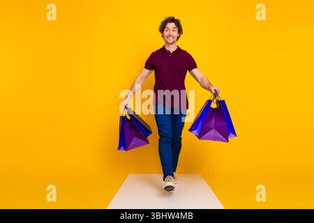 Glücklicher junger Mann mit Einkaufstaschen vor leuchtendem gelbem Hintergrund, der lässige und stilvolle Mode mit fröhlicher Atmosphäre präsentiert Stockfoto