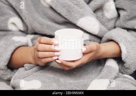 Frau im Bademantel mit Cremebecher. Gemütliches Hautpflege-Ritual zu Hause. Feuchtigkeitspflege Nahaufnahme. Sanfte Hände und Wohlbefinden. Entspannende Schönheitsbehandlung. Stockfoto