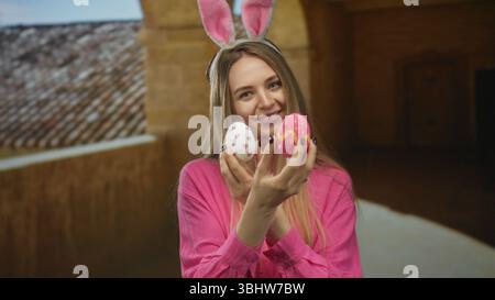 Eine junge Frau mit Hasenohren hält farbenfrohe ostereier in einem historischen Universitätsgebäude, in dem eine fröhliche, festliche Atmosphäre drinnen herrscht. Stockfoto
