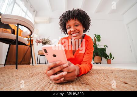 Fröhliche Frau genießt ihre Zeit, während sie auf dem Boden liegt und ihr Smartphone verwendet, um mit Freunden über verschiedene Anwendungen oder soziale Netzwerke zu kommunizieren Stockfoto