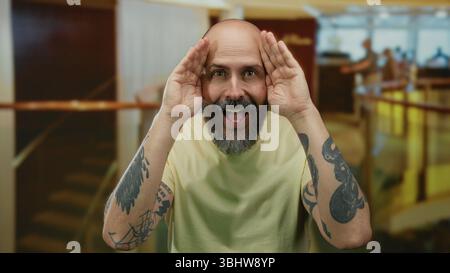 Bärtiger Mann mit Tätowierungen, der das Gesicht in einer Hotellobby verspielt bedeckt und das Charisma und die einnehmende Persönlichkeit im Innenbereich zum Ausdruck bringt. Stockfoto