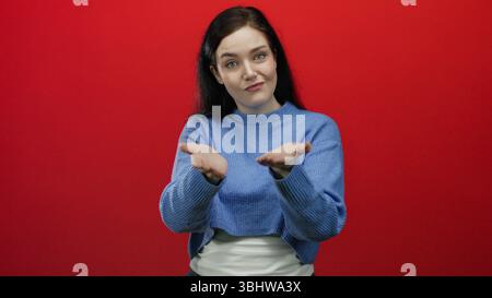 Frau in blauem Pullover gibt kreativ Gesten vor leuchtend rotem Hintergrund und drückt Eleganz und Emotionen mit brünetten Haaren und einer selbstbewussten Haltung aus Stockfoto
