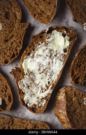 Blick von oben auf handwerklich geschnittenes Roggensauerteigbrot mit Butter aus nächster Nähe. Einfaches Frühstück auf grauem Betontisch Stockfoto