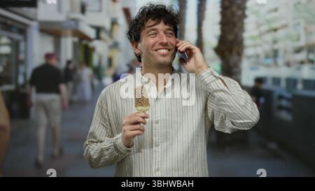 Hispanischer Mann, der Eis genießt, während er draußen auf der belebten Stadtstraße telefoniert, an einem sonnigen Tag in einem gestreiften Hemd. Stockfoto