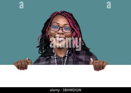 Fröhliche Afroamerikanerin mit Brille und bunten Haaren, die ein weißes Schild hält. Perfekt für Werbung, Werbeaktionen, Ankündigungen und Marketing Stockfoto
