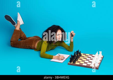 Junge Frau schreibt Notizen beim Schach auf einem leuchtend blauen Hintergrund, der Freizeit und Strategie zeigt Stockfoto
