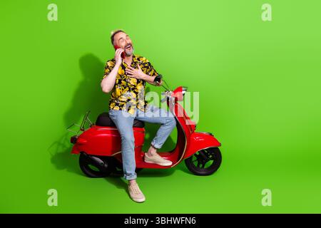 Mann mittleren Alters auf einem roten Roller mit Telefon auf grünem Hintergrund, tragend ein hawaiianisches Sommerhemd und zeigte Glück Stockfoto