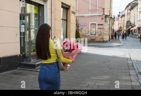 Lemberg, Ukraine - 1. Juni 2025: Eine Frau in gelbem Oberteil und Jeansrock hält einen Blumenstrauß aus roten Rosen auf einer Stadtstraße. Stockfoto