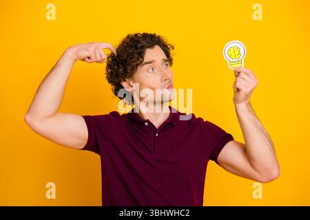 Junger Mann im lässigen burgunderfarbenen Polo, der eine Gehirnlampe vor gelbem Hintergrund hält und kreative Ideen präsentiert Stockfoto