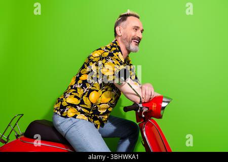 Reifer Mann mit stilvollem Bart posiert auf einem leuchtend roten Roller mit einem Hemd mit tropischem Muster vor einem üppig grünen Hintergrund Stockfoto