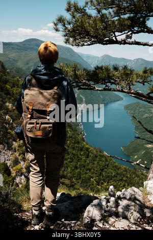 Einsame Wanderer mit Retro-Rucksack am Klippenrand des Tara-Nationalparks mit atemberaubendem Blick auf den Drina River und die grünen Berge. Hinten Stockfoto