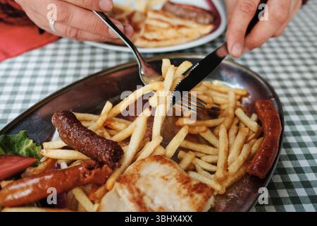 Man bemannt Hände mit Messer und Gabel, um Pommes frites mit Würstchen und Fleisch auf einem Metallteller zu essen. Serbische gemischte Grillplatte. Ansicht schließen. Balkanküche. Stockfoto