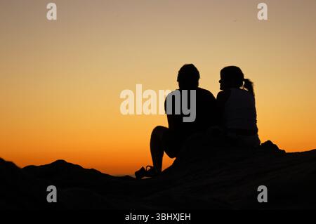 Ein junges erwachsenes Paar genießt einen romantischen Blick auf den Sonnenuntergang über dem Pazifischen Ozean von einem Barsch in Laguna Beach, Kalifornien Stockfoto