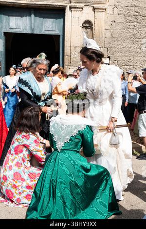 Die Königliche Königin in weiß steht vor der Kirche, während zwei Frauen ihr langes Kleid zu Beginn des Fete du Kostüm Festivals anpassen, wo über 50 Stockfoto