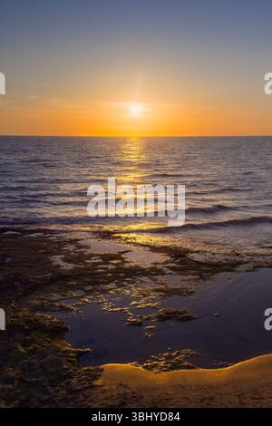 Goldenes Licht tanzt über dem Wasser, während Wellen sanft gegen eine ruhige Küste bei Sonnenuntergang schlagen. Stockfoto