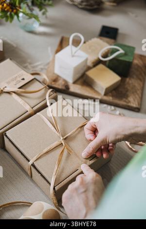Person bindet Band um Kraftgeschenkboxen auf einem Tisch mit handgefertigten Seifen und Verpackungsmaterialien. Kleinserienproduktion, handwerkliche Wirtschaft, Hersteller Stockfoto