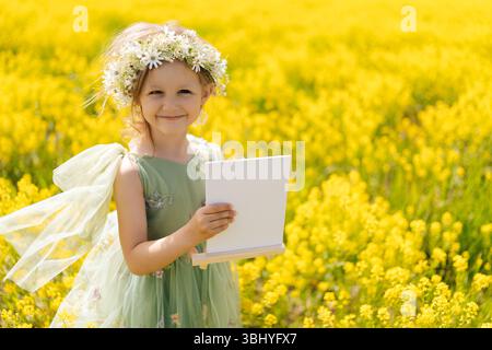 Hübsches Mädchen mit Blumenkrone und Feenkleid, das leere Leinwand auf hölzerner Staffelei hält und auf grüner Wiese voller gelber Blumen posiert Stockfoto