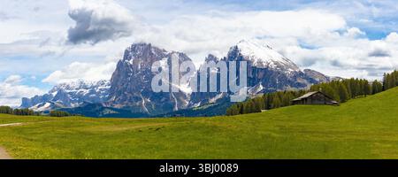 Panoramablick auf die majestätische schneebedeckte Langkofel-Berggruppe von den riesigen grünen Wiesen der Seiser Alm, mit einer traditionellen Almhütte. Stockfoto