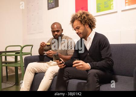 Zwei Profis arbeiten in einem Coworking-Bereich in Barcelona. Sie sitzen auf einem gemütlichen Sofa, unterhalten sich und arbeiten zusammen, halten Kaffeetassen in der Hand, mit lebendigem DE Stockfoto
