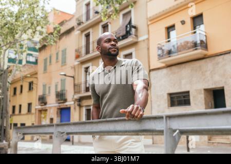 Ein Mann genießt einen entspannten Moment außerhalb eines Coworking-Raums in Barcelona. Er lehnt sich beiläufig auf ein Geländer, umgeben von charmanten alten Gebäuden und Baum-l Stockfoto