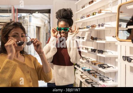 Zwei Freunde erkunden einen stilvollen Sonnenbrillenladen und probieren verschiedene Paare an. Die Regale sind mit verschiedenen Designs gefüttert und sorgen so für lebendige und ansprechende s Stockfoto