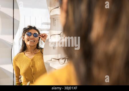 Eine lächelnde Frau in einem Senfpullover probiert eine stilvolle Sonnenbrille an, während sie in einem modernen Geschäft im Spiegel schaut. In den Regalen werden verschiedene Rahmen in der Ba angezeigt Stockfoto